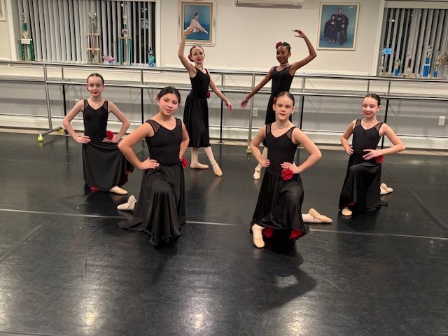 Five dancers in black and red costumes pose in a studio, with two standing behind and three kneeling in front.