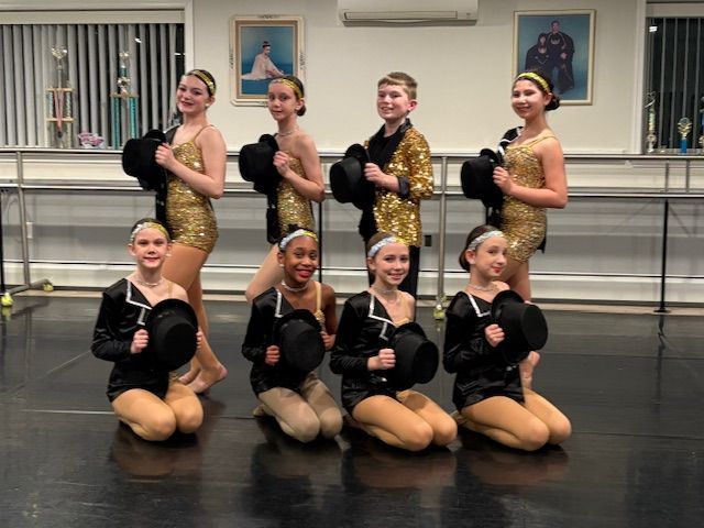 Eight dancers in black and gold costumes pose in a studio with ballet barres and framed art.