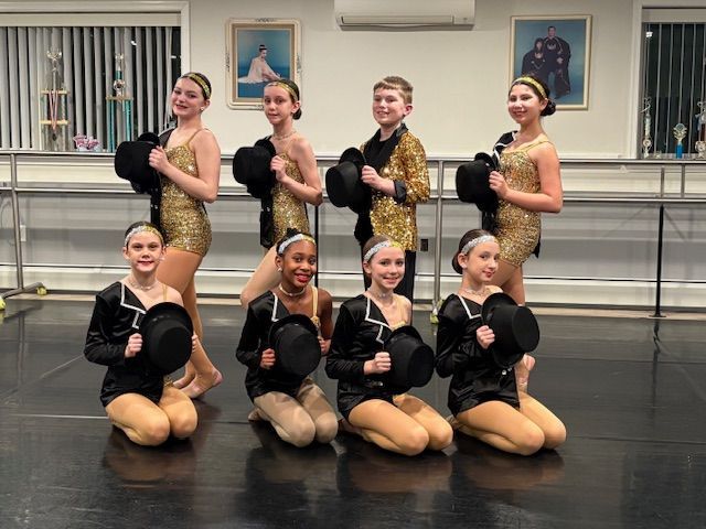 Eight dancers in black and gold costumes pose in a studio with ballet barres and framed wall art.