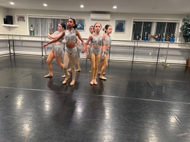 Four dancers in silver costumes pose in a dance studio with mirrors and black flooring.