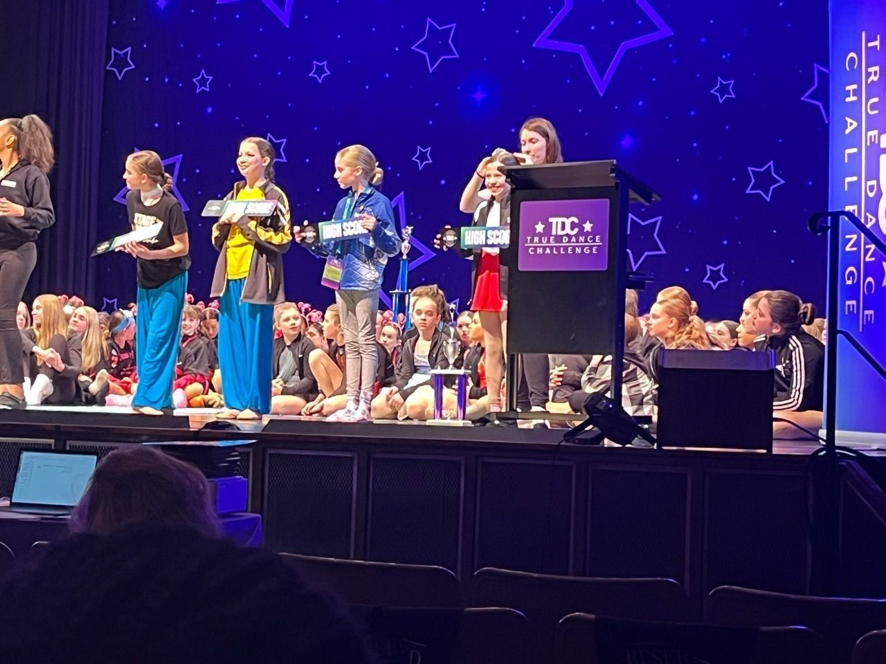 Children on a lit stage receiving awards, with a podium and seated audience in the background.