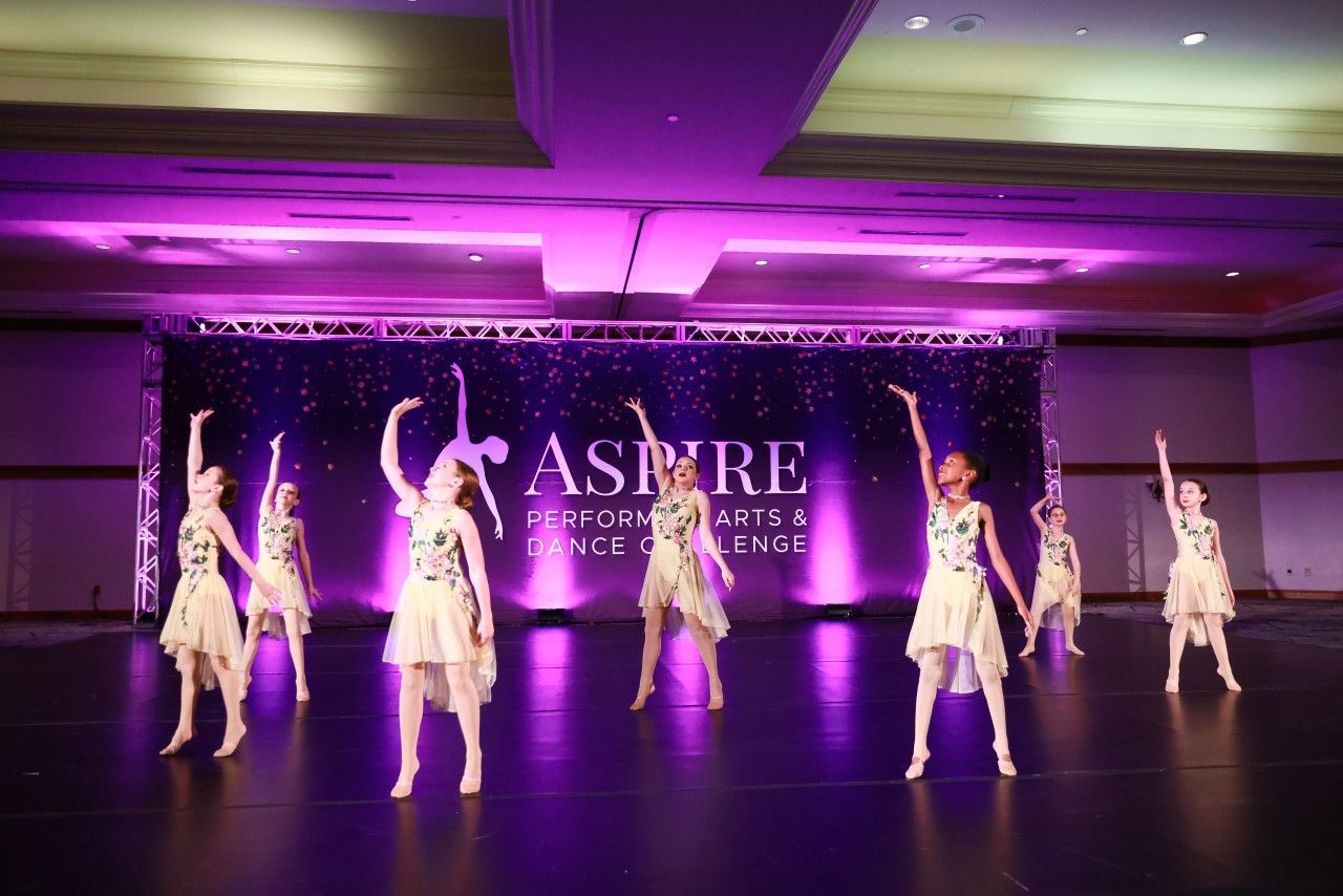 Six dancers in white costumes performing on a purple-lit stage in a ballroom with an ASPIRE backdrop
