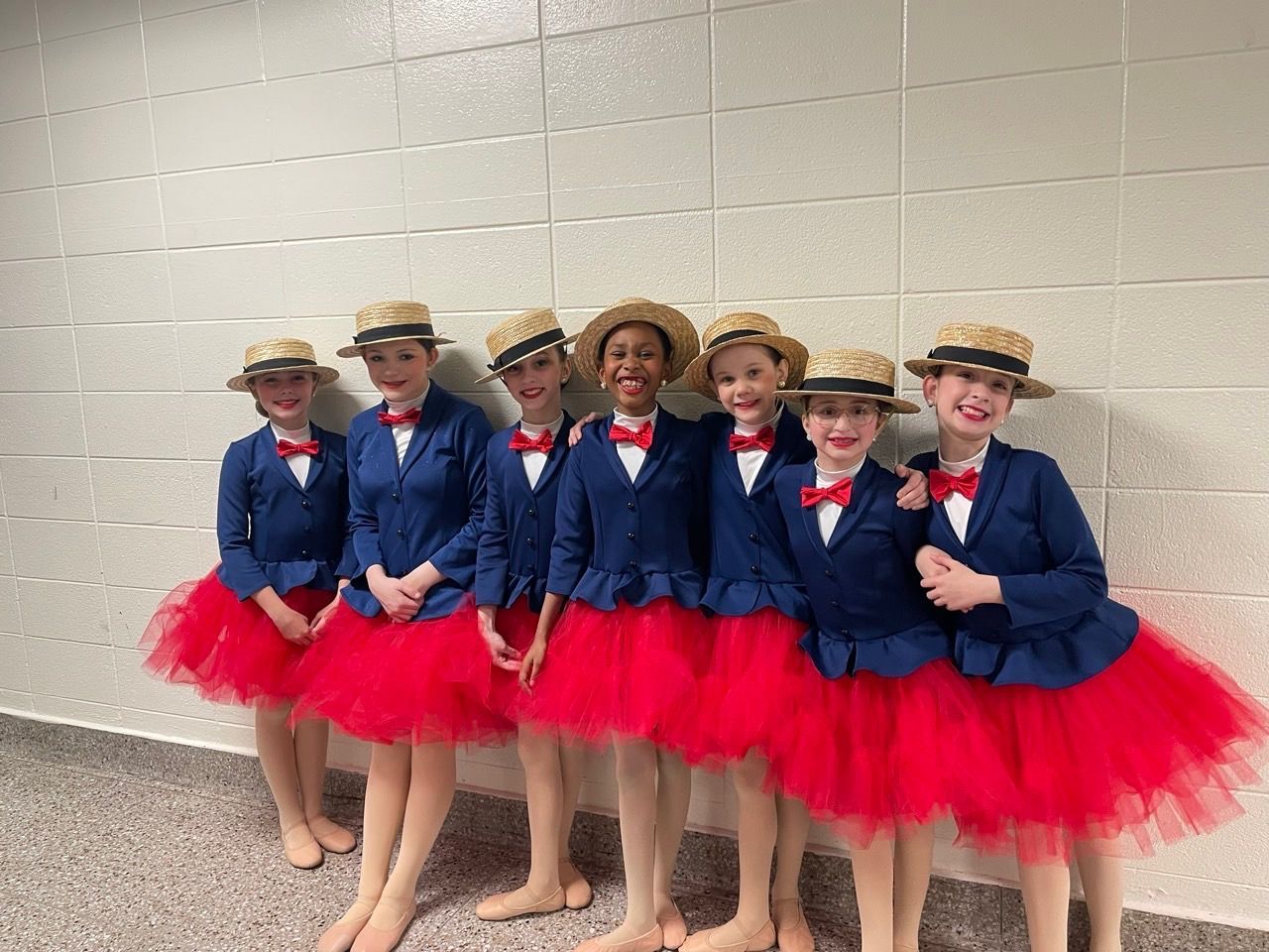 Six children in matching blue jackets, straw hats, and red tutus posing against a wall.