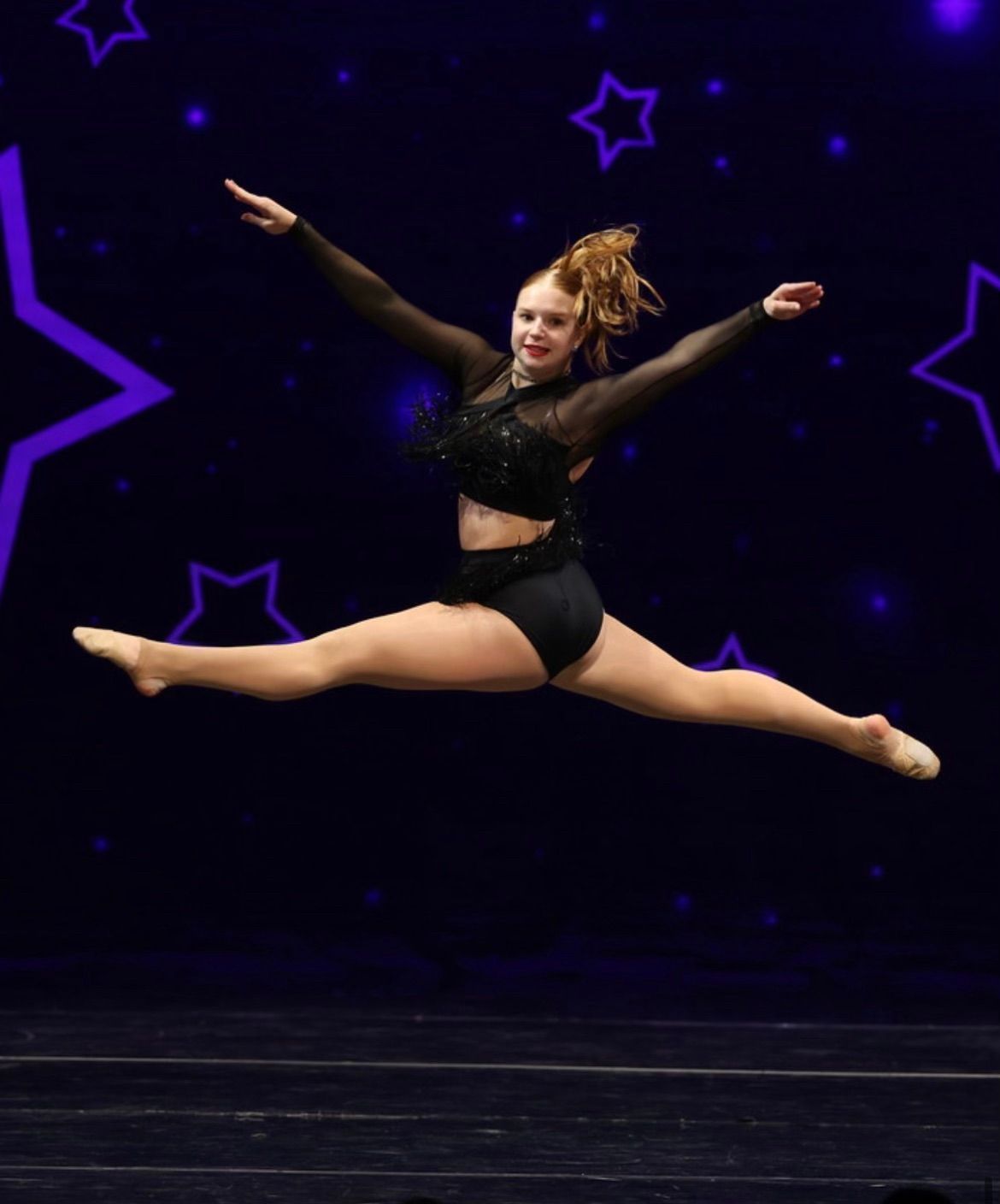 Dancer in a black costume mid-air in a split leap against a dark stage with purple star lights