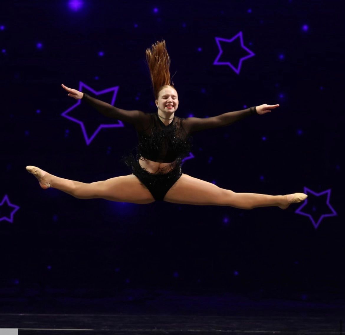 Dancer in black costume doing a split leap on a dark stage with purple star lights