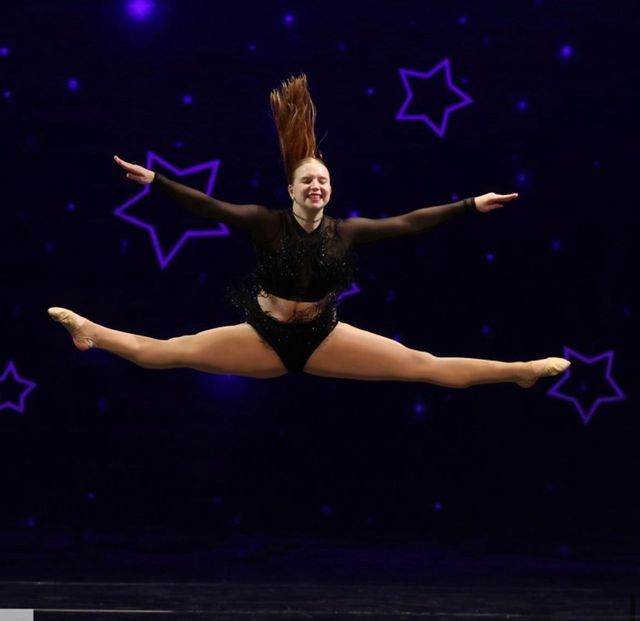 Dancer in a black outfit performing a split leap on a dark stage with purple star outlines