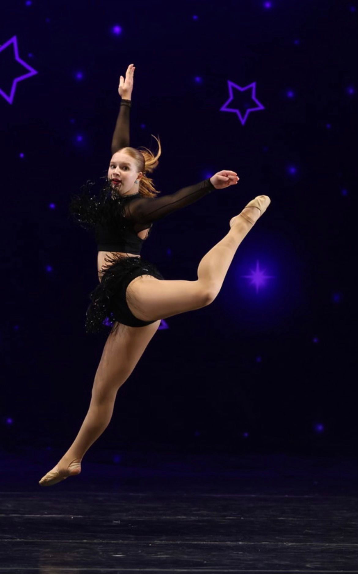 Dancer in black costume performing a high leg extension on a dark stage with purple star accents