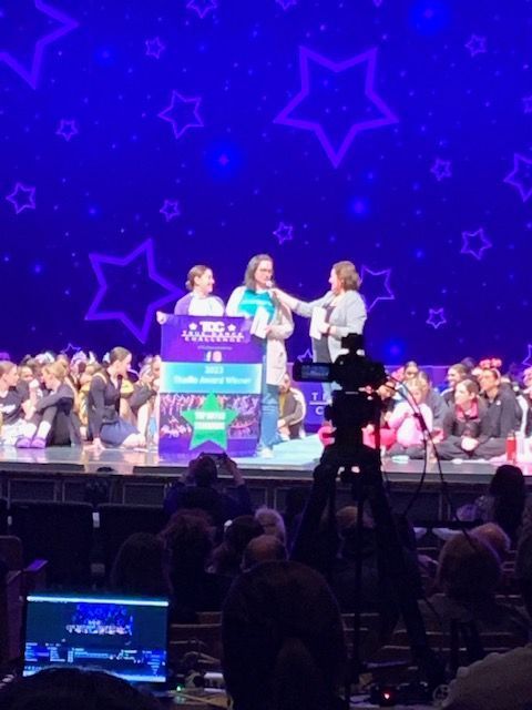 Three people onstage at a purple-lit event, standing behind a podium with a cheering crowd and starry backdrop.