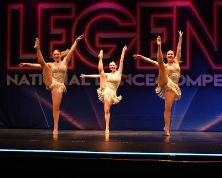 Three dancers in gold costumes perform onstage in front of a neon “LEGEND” backdrop.
