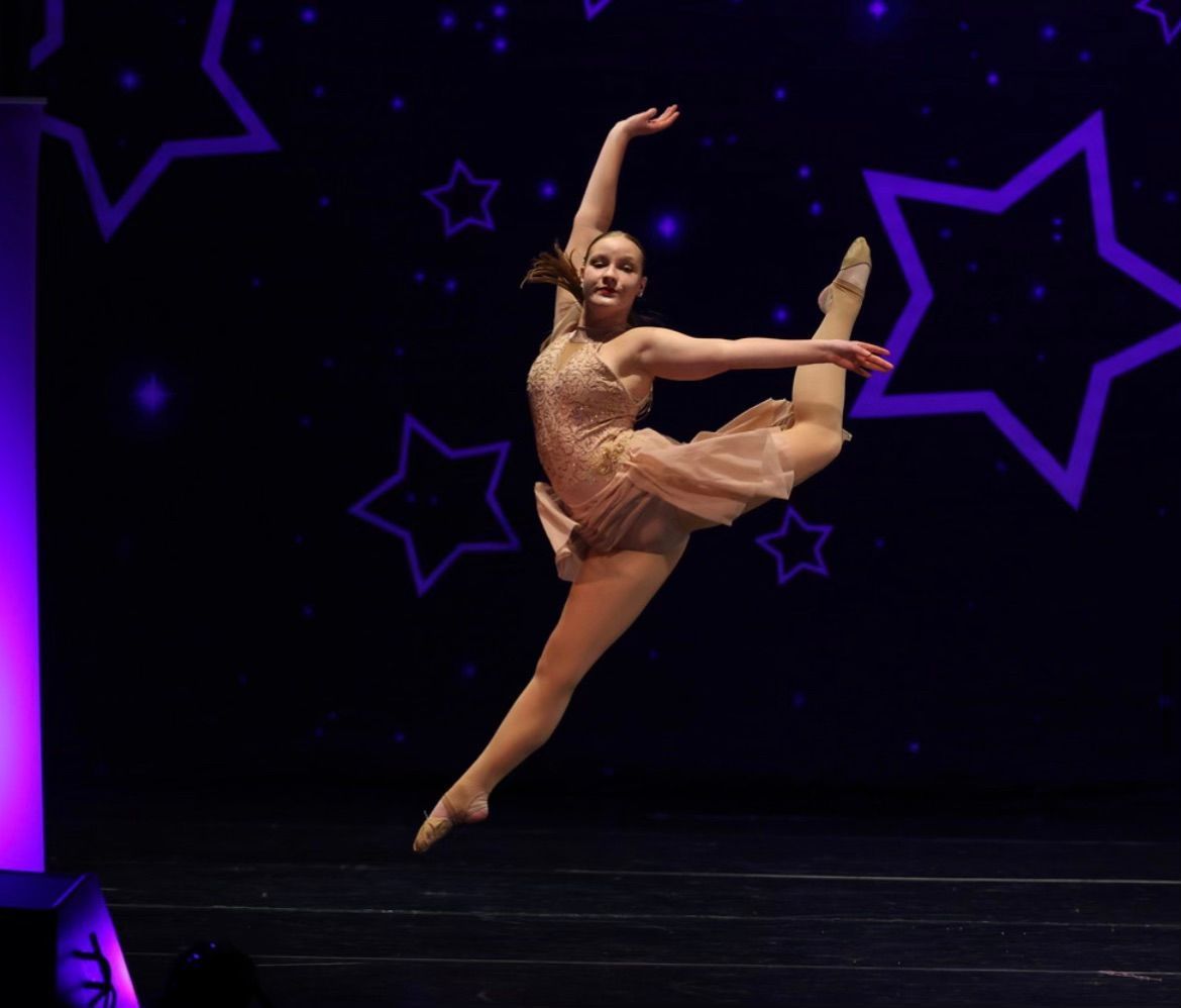 Dancer in a beige costume performs a high leg extension on a dark stage with purple star-shaped lights