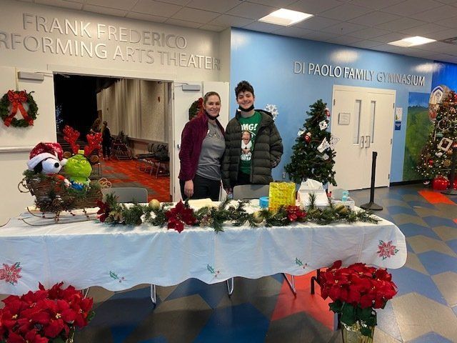 Two people behind a decorated holiday display table in a lobby with a wreath, poinsettias, and small Christmas trees.