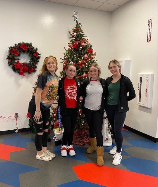 Four people pose in front of a Christmas tree and wreath in a decorated indoor hallway.