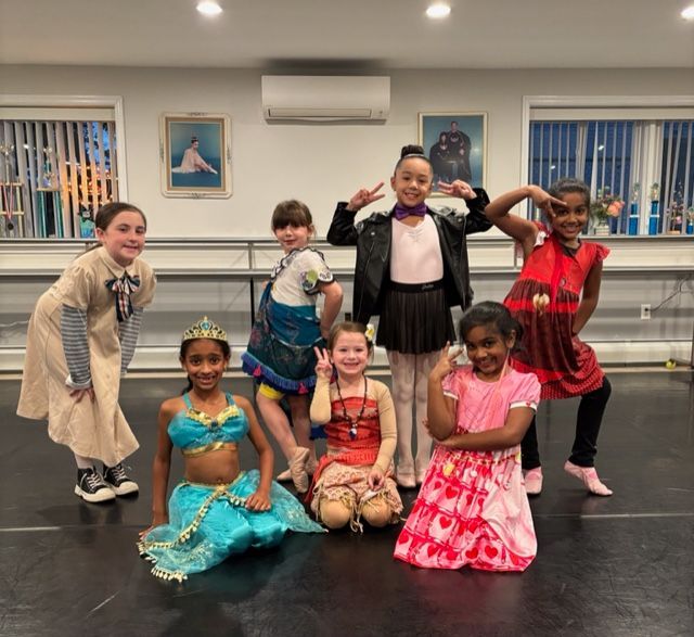 Seven children in colorful costumes pose in a dance studio, smiling and striking playful poses.