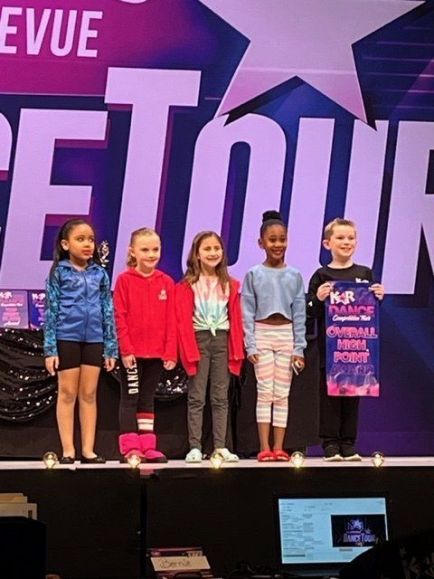 Five children stand on a stage holding a large purple event sign, with a “Revue” backdrop behind them.