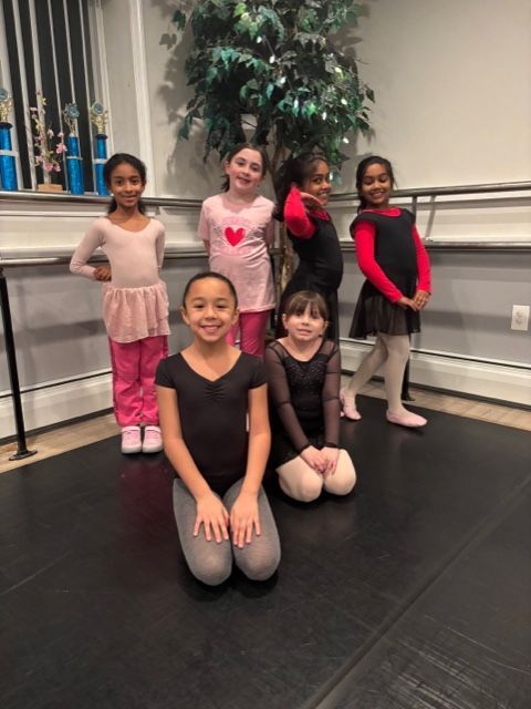 Five children in ballet outfits posing in a dance studio, with trophies and a plant in the background