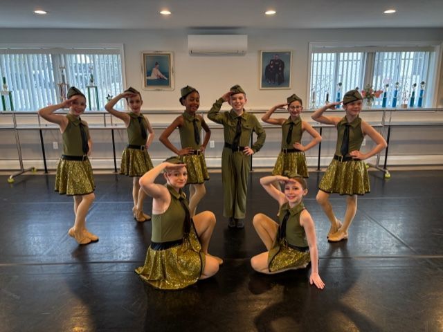 Seven dancers pose in a studio, wearing olive and gold costumes with one kneeling in front.