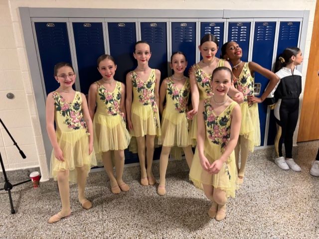 Seven dancers in yellow costumes pose in a hallway with blue lockers.
