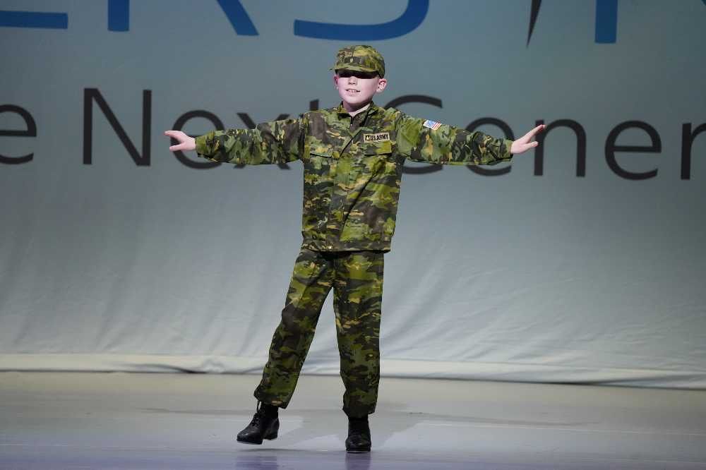 Person in camouflage uniform stands on stage with arms outstretched before a large backdrop.