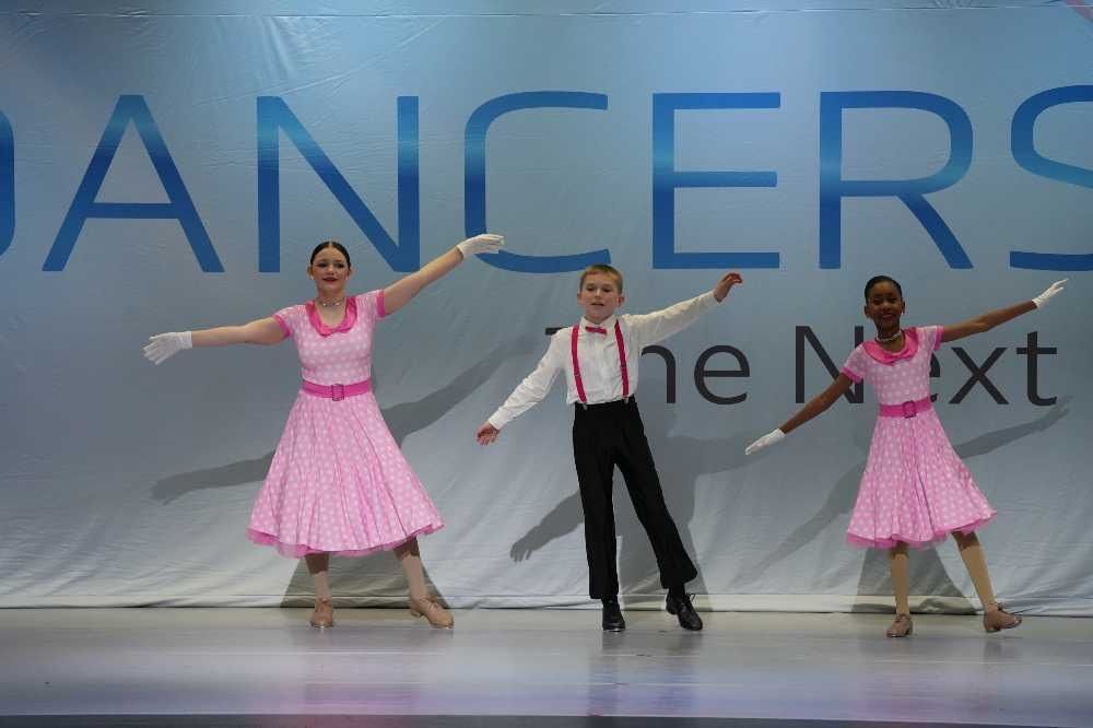 Three dancers perform onstage; two in pink dresses flank a dancer in a black suit with red tie.