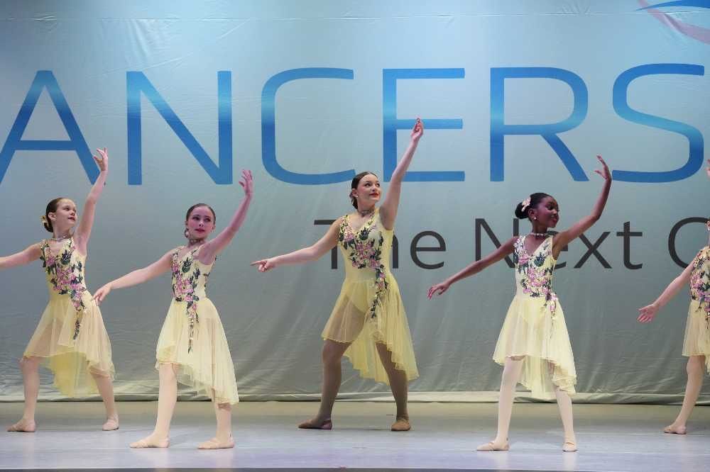 Five dancers in yellow costumes perform on a stage with a large “DANCERS” backdrop.