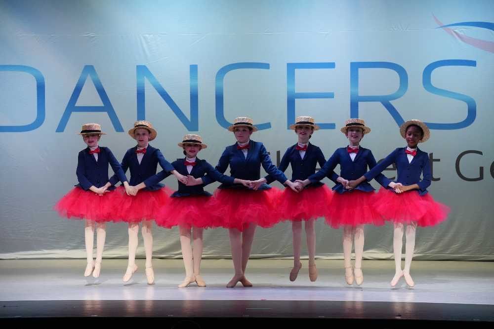 Six dancers in navy and pink costumes stand in a line on a stage with a “DANCERS” backdrop.