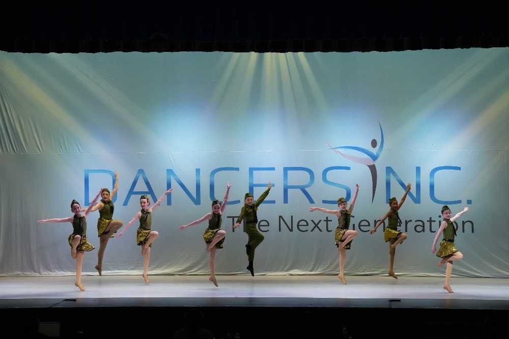 Five dancers perform on a stage with a blue backdrop reading “DANCERS INC” and “The Next Generation.”