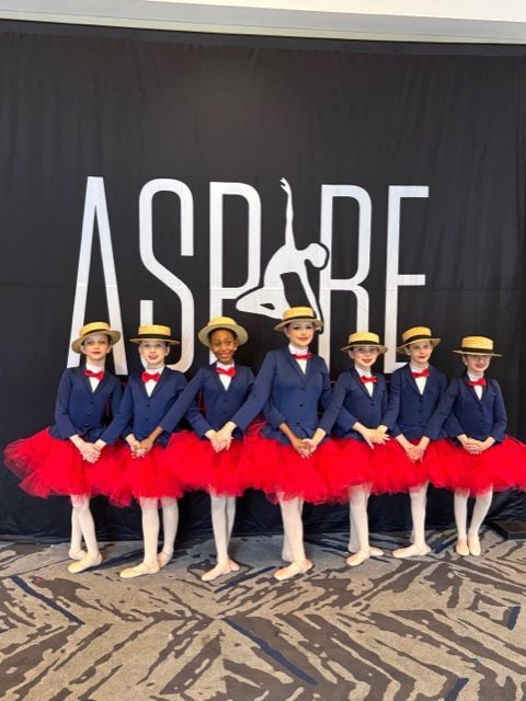 Seven dancers in blue jackets and red tutus posing onstage before an ASPIRE backdrop.