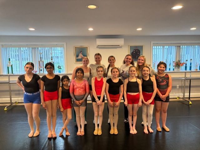 Group of dancers posing in a studio, wearing black, red, and blue leotards with ballet shoes.