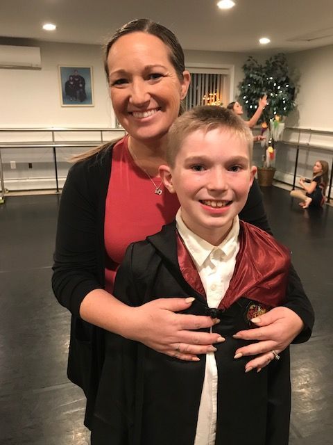 Two smiling people posing indoors, one in a black and red costume, with dance studio mirrors in the background