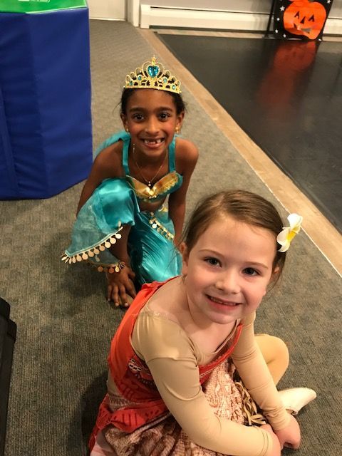 Two children in colorful princess costumes kneeling indoors, smiling at the camera.