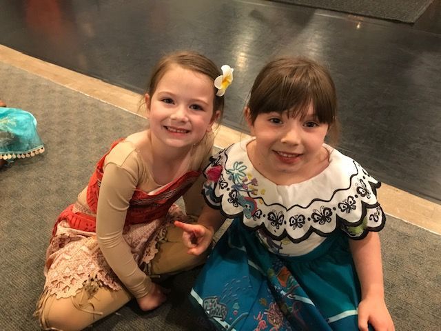 Two children sitting on a carpet, smiling at the camera, in colorful dresses.