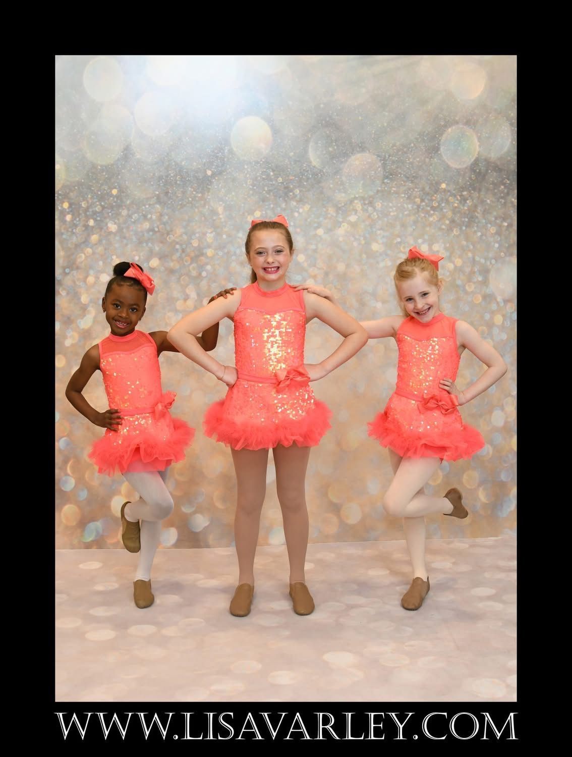 Three young dancers in coral tutus pose on tiptoe against a sparkly backdrop.