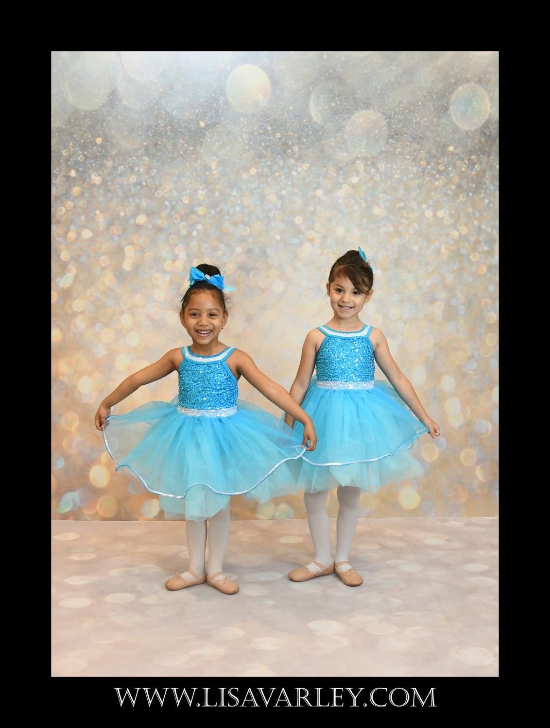 Two young ballet dancers in blue tutus holding hands against a sparkly studio backdrop