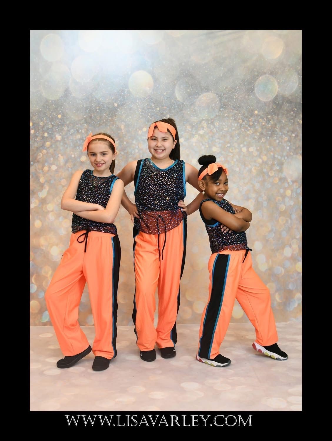 Three dancers in orange pants and sequined tops pose against a sparkly backdrop.