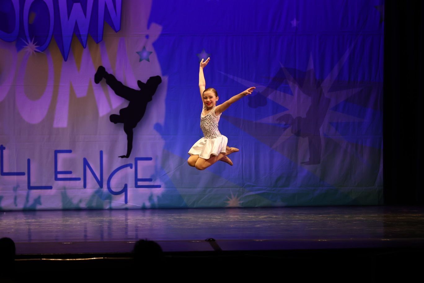 Dancer mid-leap on a purple-lit stage with a shadow silhouette in the background