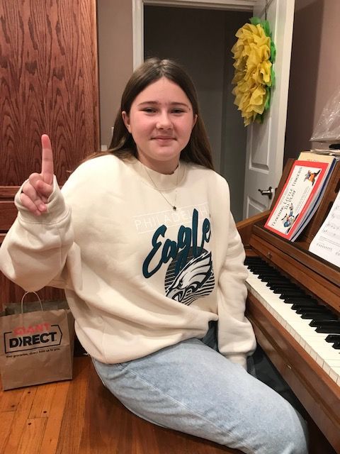 Person seated at a piano, raising one finger, wearing a white sweatshirt and jeans in a home music room