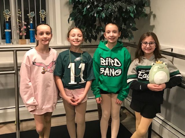 Four kids posing indoors by a window, wearing sports-themed shirts; one holds a white plush ball.