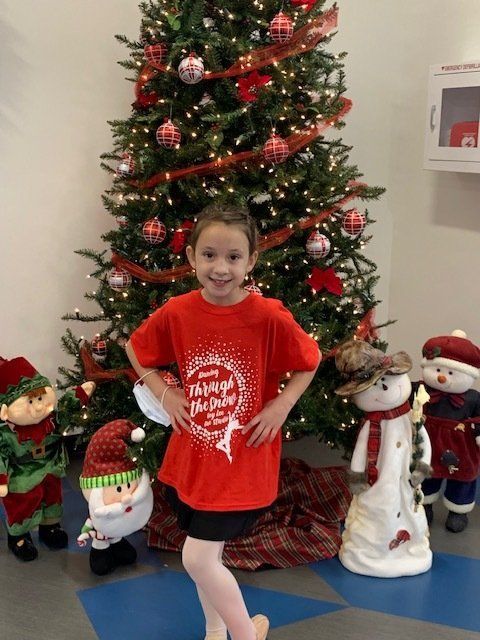 Child in red shirt posing in front of a decorated Christmas tree with holiday figures.