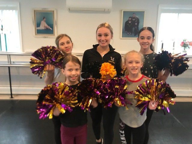 Five dancers holding pom-poms in a studio, smiling in front of ballet barres and framed portraits.