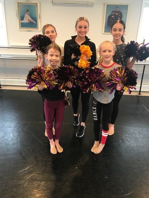 Five dancers posing in a studio with purple and orange pom-poms, smiling in front of a mirror