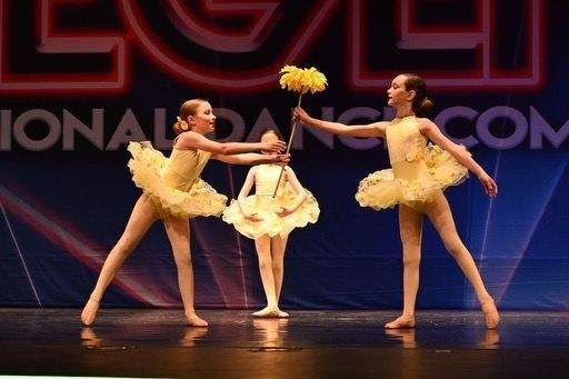 Three ballet dancers in yellow tutus pose on stage with a large red and blue backdrop.
