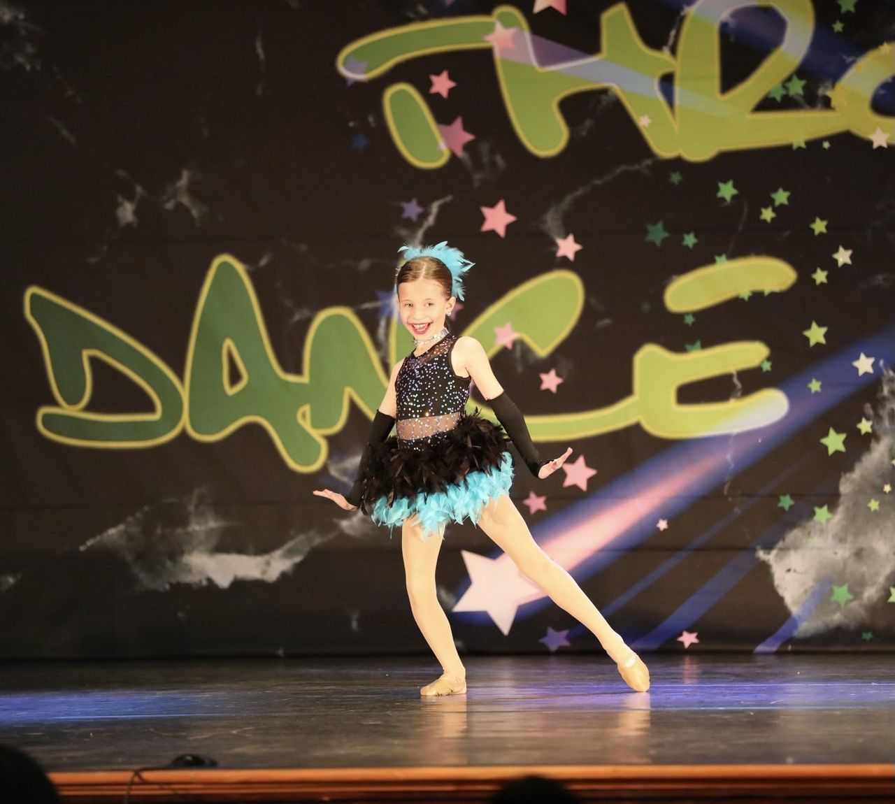 Young dancer in black and blue costume performing on a stage with graffiti-style backdrop and pink stars