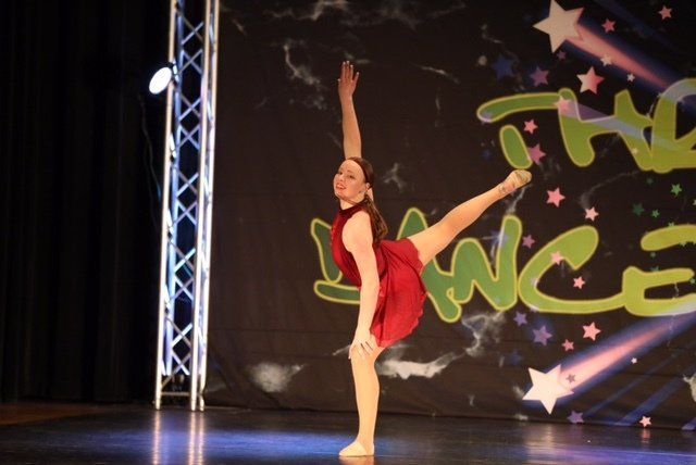 Dancer in red costume balancing on one leg with arms raised on a stage with starry backdrop