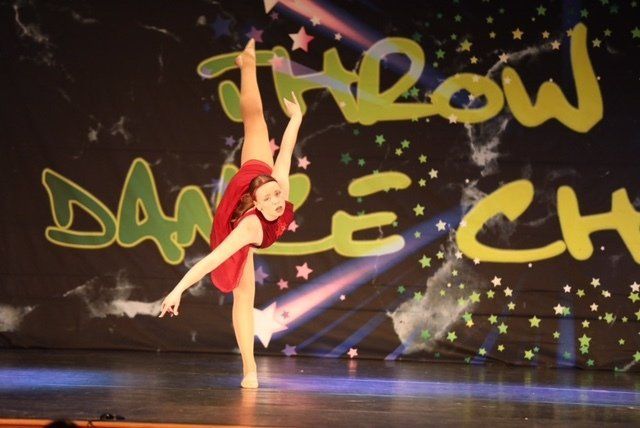 Dancer in red leotard performs a high leg extension on stage before a 