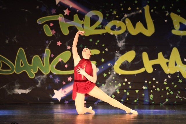 Dancer in a red costume posing onstage with a raised arm against a colorful dance backdrop