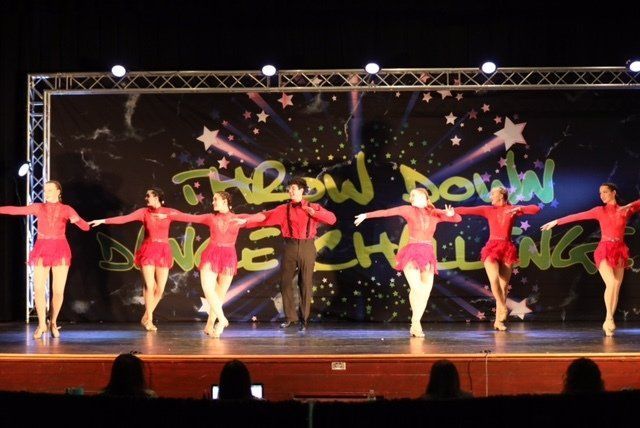 Six dancers in red costumes perform on stage under “THE SHOW MUST GO ON” backdrop.