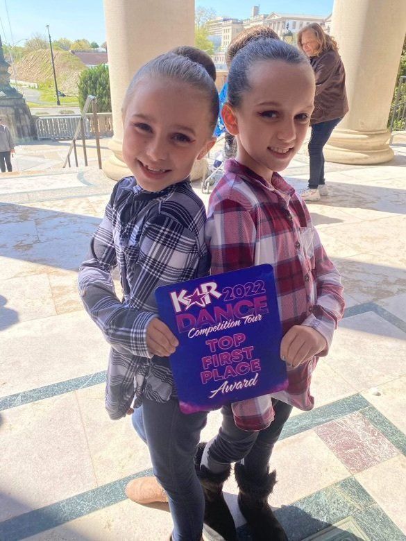 Two children standing back to back, smiling and holding a purple sign outdoors in bright sunlight.