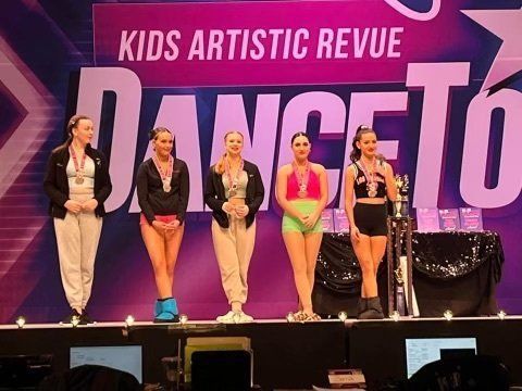 Five performers stand onstage under a “Kids Artistic Revue” banner during an awards event.