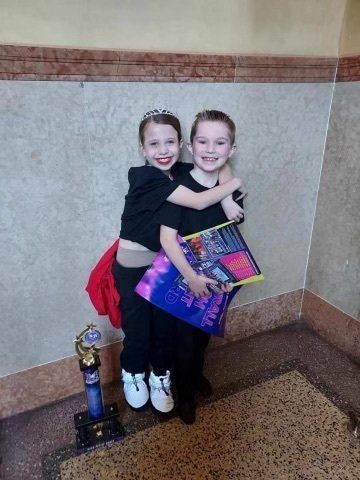 Two smiling children posing together indoors, one holding a colorful gift bag and trophy, beside a wall.