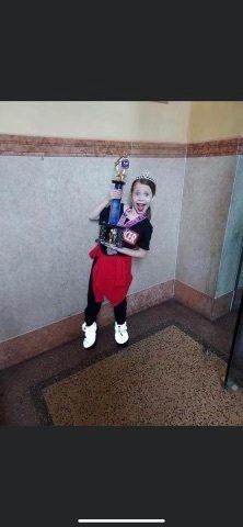 Child holding a doll, standing against a hallway wall in a red outfit and white shoes.
